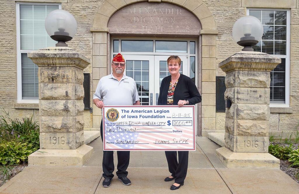 A person with an American legion cap, Dennis Soppe, presents a large check with an American flag background to UIU President Kathy Franken on the right.
