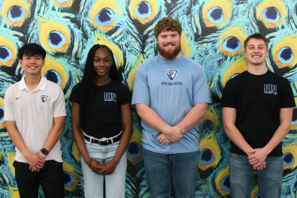 The 2026–27 SGA Council poses in front of a colorful peacock-feather mural on campus, with four student leaders standing side by side and smiling at the camera.