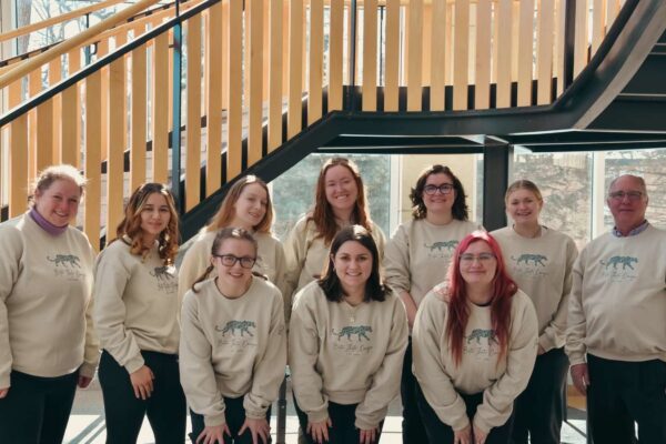Nine people in matching beige "Beta Theta Omega" sweatshirts pose, smiling, in front of a staircase and large windows.