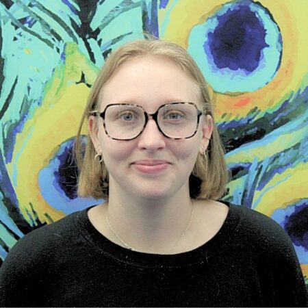 The Student Life Engagement Coordinator Memphis Dunsdon. She has a blonde bob, with black square glasses and a black long sleeve shirt. Memphis is standing in front of a peacock mural.