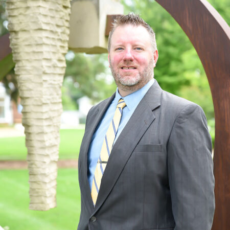 A photo of Seth Miner outside with a modern wood and stone sculpture in the background