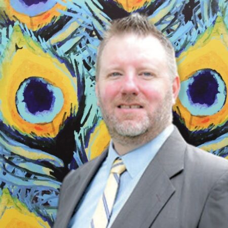 Vice President of Student Life and Admissions Seth Miner. He is wearing a grey suit, with a blue undershirt, and a tie that is white with thin blue stripes. He has short blonde hair.