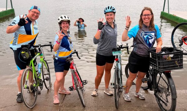 Team Peacock riders standing in front of the Mississippi River holding up a peacock symbol with their bikes.
