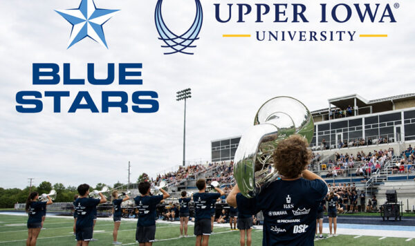 A low angle of a marching band in formation with a few up to a crowd of spectators. The Blue Stars and Upper Iowa University logos are present