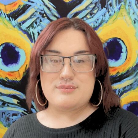 Residence Area Coordinator Zachary Z Hoskins is wearing a black sweater, with thin rimmed square glasses. Their hair is red, long, and hanging loose. Standing in front of a peacock mural.