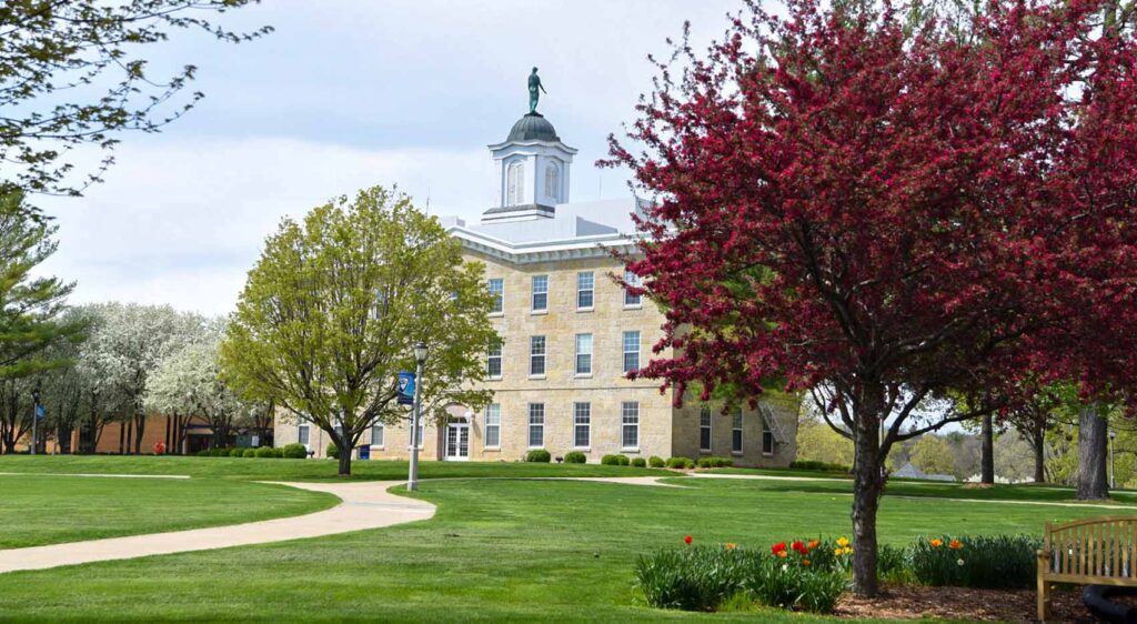 A large, historic limestone building in the background, surrounded by a lush green lawn, and colorful blossoming trees and tulips in the foreground
