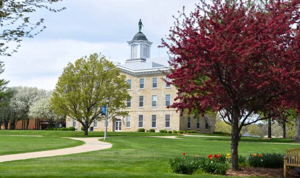A large, historic limestone building in the background, surrounded by a lush green lawn, and colorful blossoming trees and tulips in the foreground