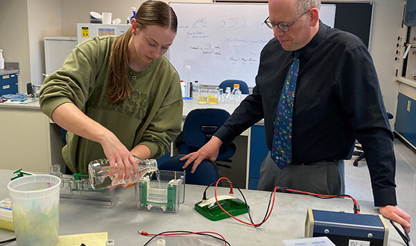 A student pours a liquid into a container under the supervision of a professor in a classroom laboratory setting.