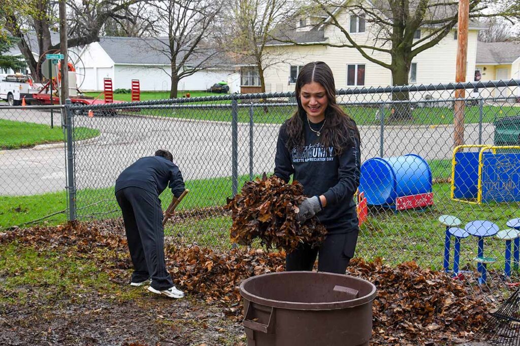 Two UIU students are shown cleaning up leaves near a playground on a rainy day