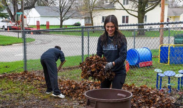 Two UIU students are shown cleaning up leaves near a playground on a rainy day