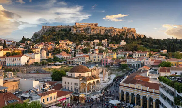A wide-angle view of Athens, Greece, with ancient ruins on a hilltop in the background. Picturesque cream-colored buildings with terra-cotta colored roofing are in the foreground.