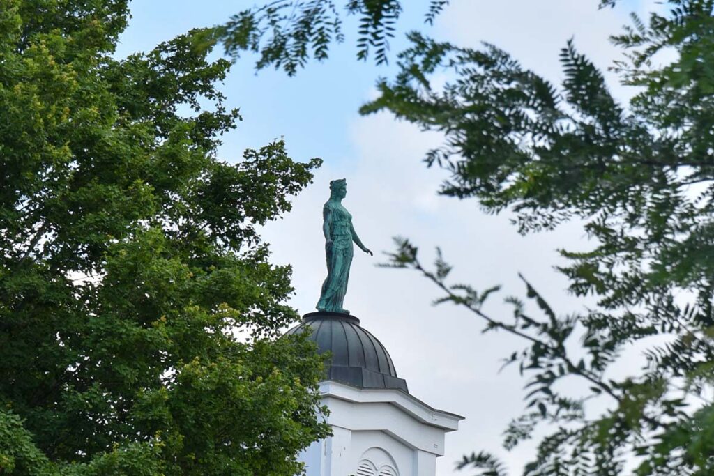 A view between two tress of a copper goddess statue with green patina sits on top of a domed structure.