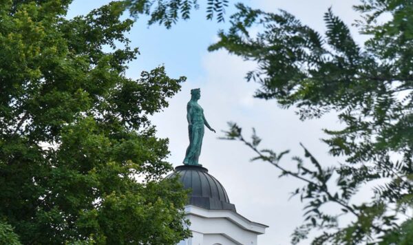 A view between two tress of a copper goddess statue with green patina sits on top of a domed structure.