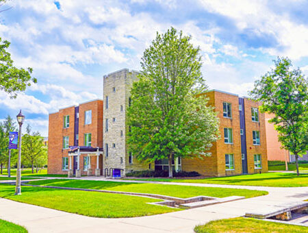 A large building with orange and sand colored bricks, with a large central "tower" section in the middle, with a large green tree and fresh mowed grass out front.