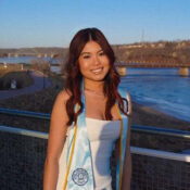 A woman in a white dress poses with their light-blue UIU graduation sash and cap with a river and bridges in the background.