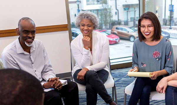 A group of people sit in a circle during a counseling session. One person has a legal pad.