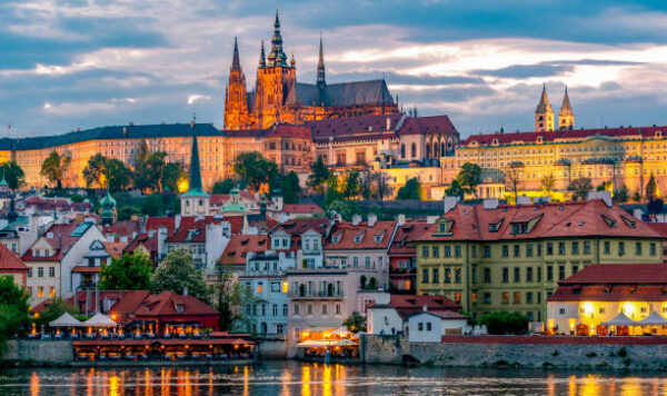A picturesque view of an an old city at sunset, from the viewpoint of a river. Colorful buildings an a large church are depicted.
