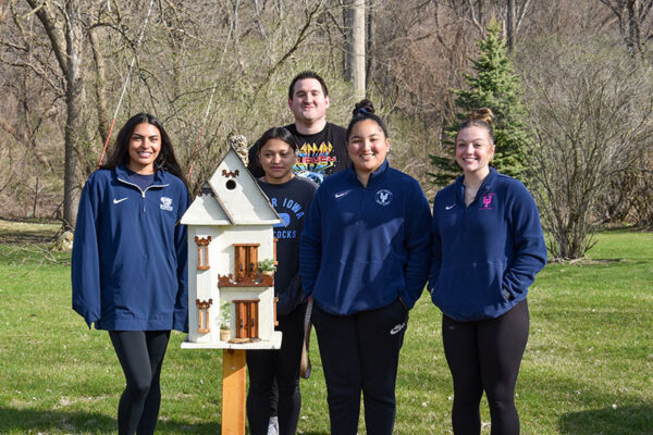 A group of students, three in matching UIU navy jackets, pose with a beautiful birdhouse on a sunny day