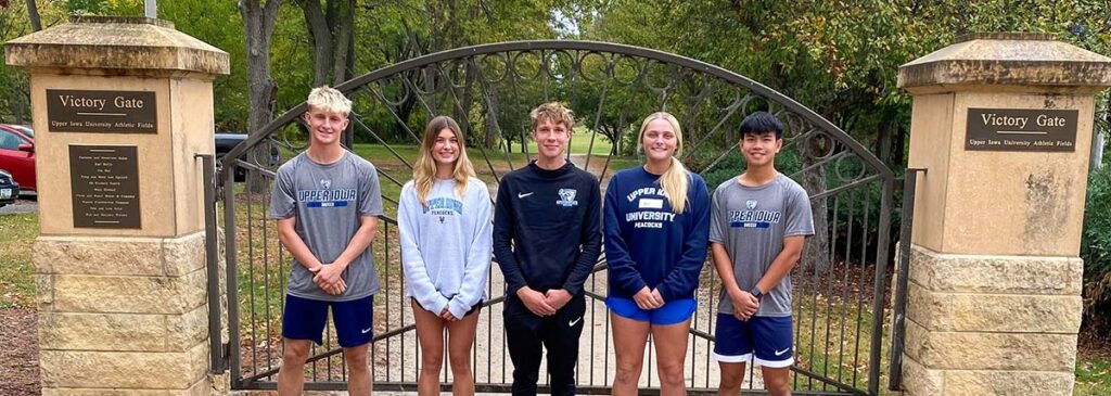 A group of UIU students pose for a group photo in front of a iron gate set between stone columns. On one column, there's a plaque that reads "Victory Gate"