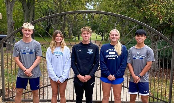 A group of UIU students pose for a group photo in front of a iron gate set between stone columns. On one column, there's a plaque that reads "Victory Gate"