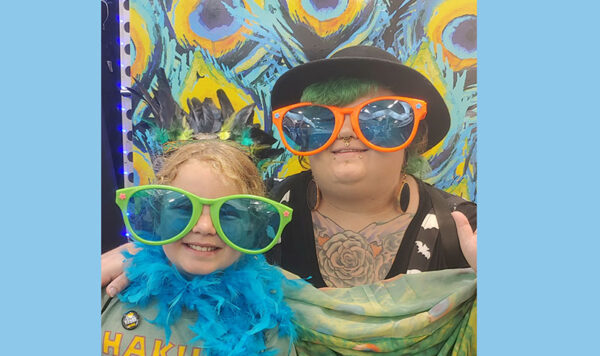 Two UIU fans with oversized colorful sunglasses pose for a photo in front of a colorful backdrop at the Iowa State Fair