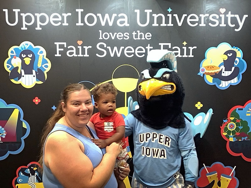 An adult and baby pose with Pete, UIU's blue Peacock mascot, at UIU's Iowa State Fair Photo Booth