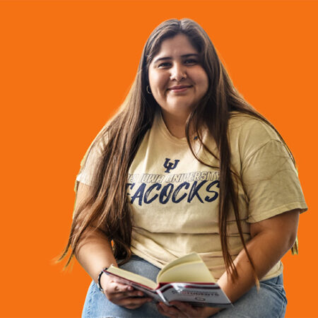 A UIU student with long hair and light yellow shirt reads a book and poses for the camera with a smile