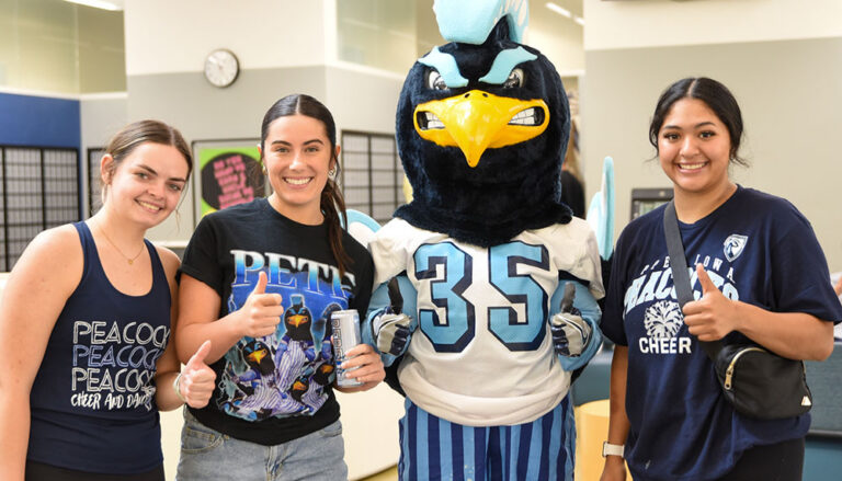 Three students wearing UIU peacock shirts pose with Pete, Upper Iowa's Peacock mascot