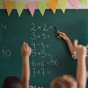A teacher's hand points to a math problem on a green chalkboard while students raise their hands