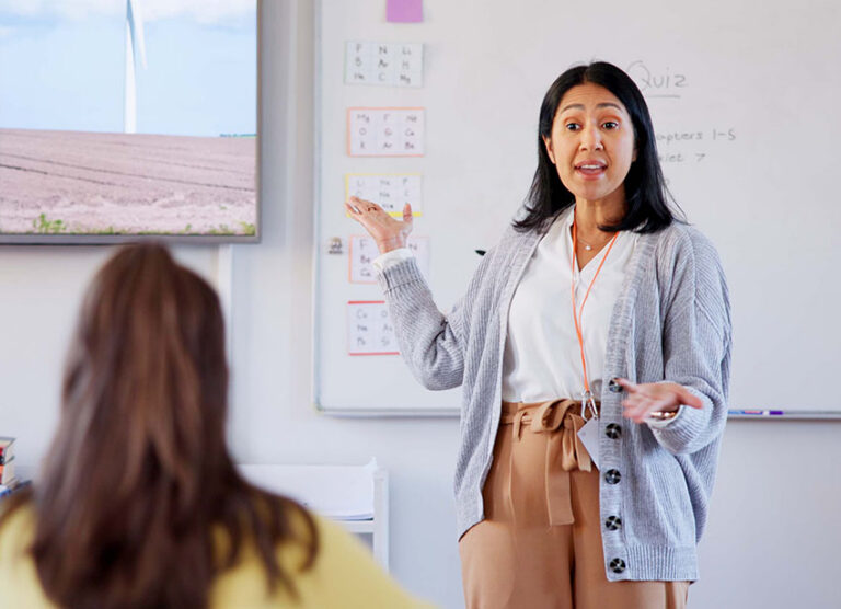 A teacher in a grey sweater presents in front of a whiteboard
