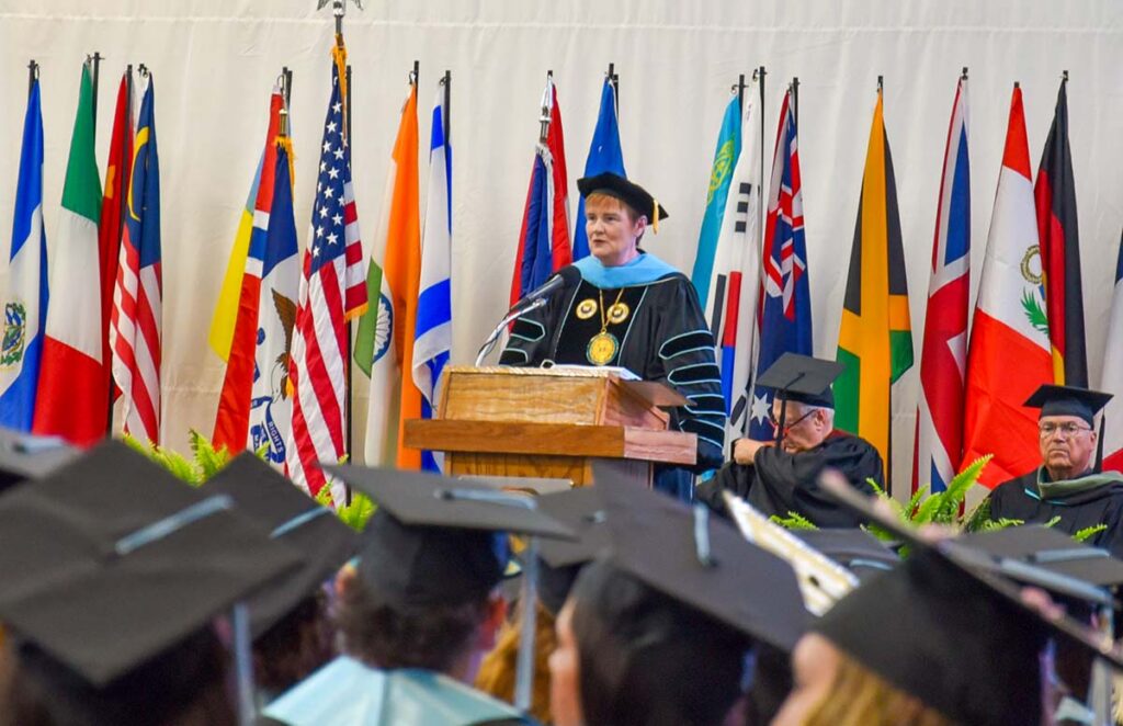 UIU's President, Kathy Franken, addresses a crowd of graduates in cap and gown from a podium with several international flags in the background.