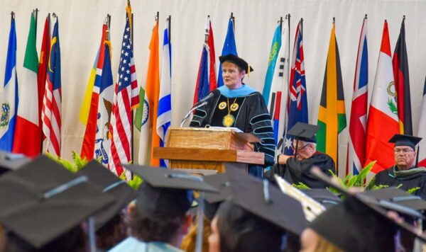 UIU's President, Kathy Franken, addresses a crowd of graduates in cap and gown from a podium with several international flags in the background.