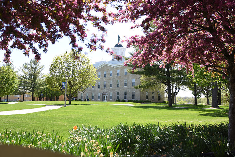 Photo of UIU's Alexander Dickman Building with blossoming trees and a green lawn in the foreground