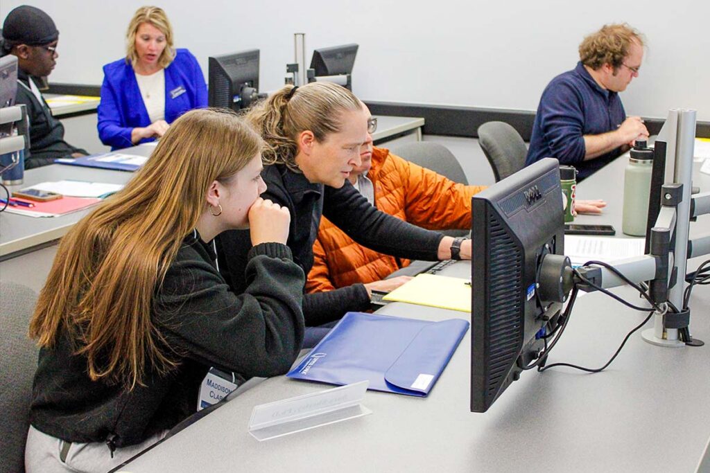 A classroom where a faculty member operates a computer between two students. A student works in the background by themself, while another faculty member discusses an item with a student in the background.