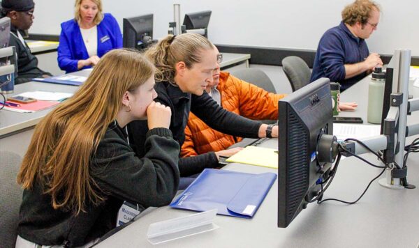 A classroom where a faculty member operates a computer between two students. A student works in the background by themself, while another faculty member discusses an item with a student in the background.