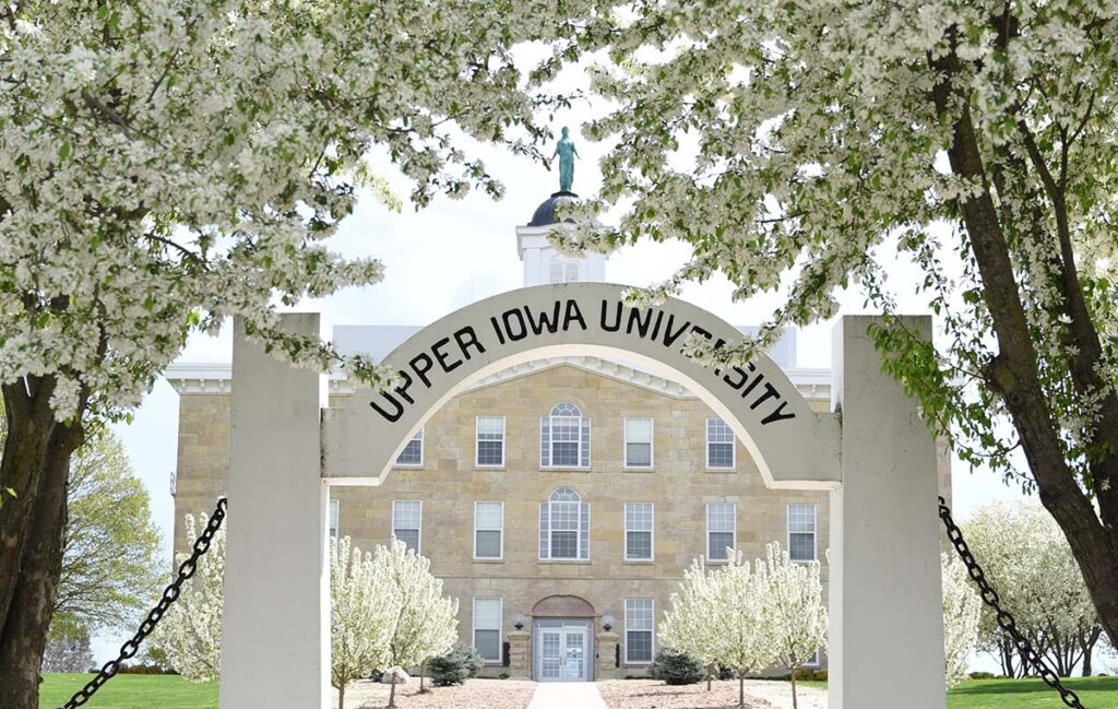 Beautiful blossoms form in the trees above an arch reading "Upper Iowa University" in the spring on UIU's Fayette, IA campus, with the historic Alexandar-Dickman building in the background.