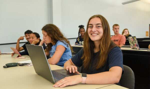 Student smiling while working on a laptop in a business classroom, with other students seated at desks in the background.