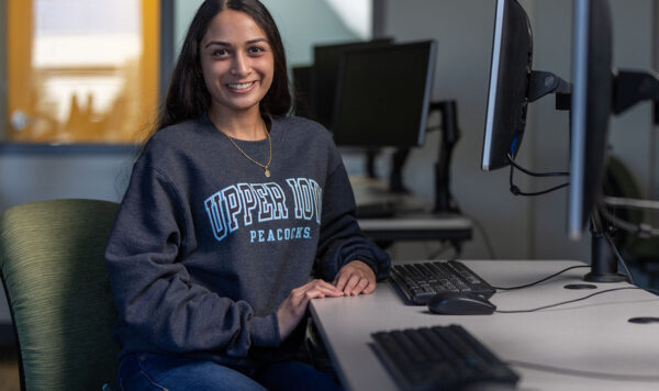A student wearing an Upper Iowa University sweater sits at a computer workstation and smiles